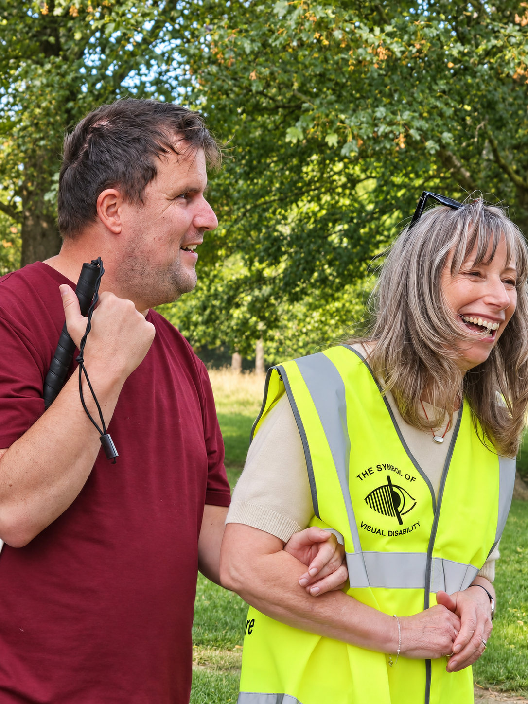 A man carrying a white cane walking arm in arm with a smiling woman in a yellow visual impairment awareness vest.